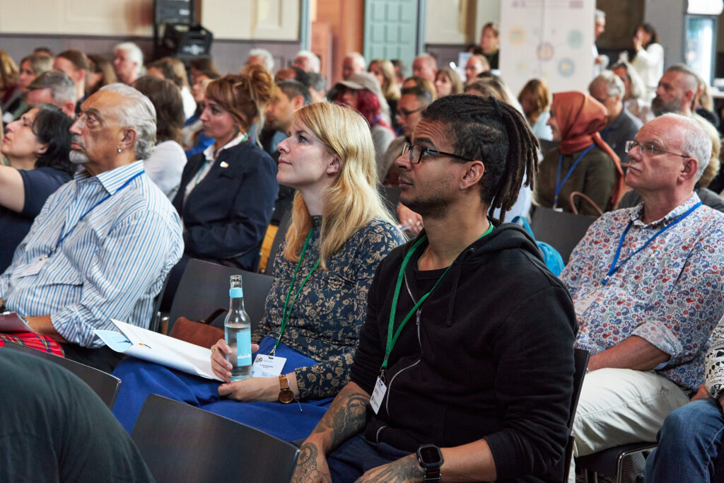Mehrere Personen sitzen im Publikum und hören aufmerksam einem Vortrag zu. Sie tragen Namensschilder und blicken nach vorn.