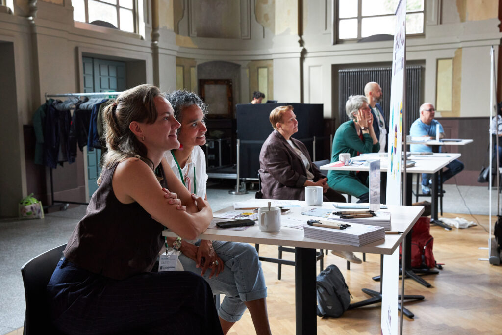 Annik Schepp und Laura Schrader von FAMAD sitzen lächelnd an einem Tisch und blicken nach vorn. Sie nehmen an einer Veranstaltung in einem großen Saal teil. Auf dem Tisch liegen Kaffeetassen, Stifte und Broschüren. Im Hintergrund sitzen weitere Personen aufmerksam an Tischen.