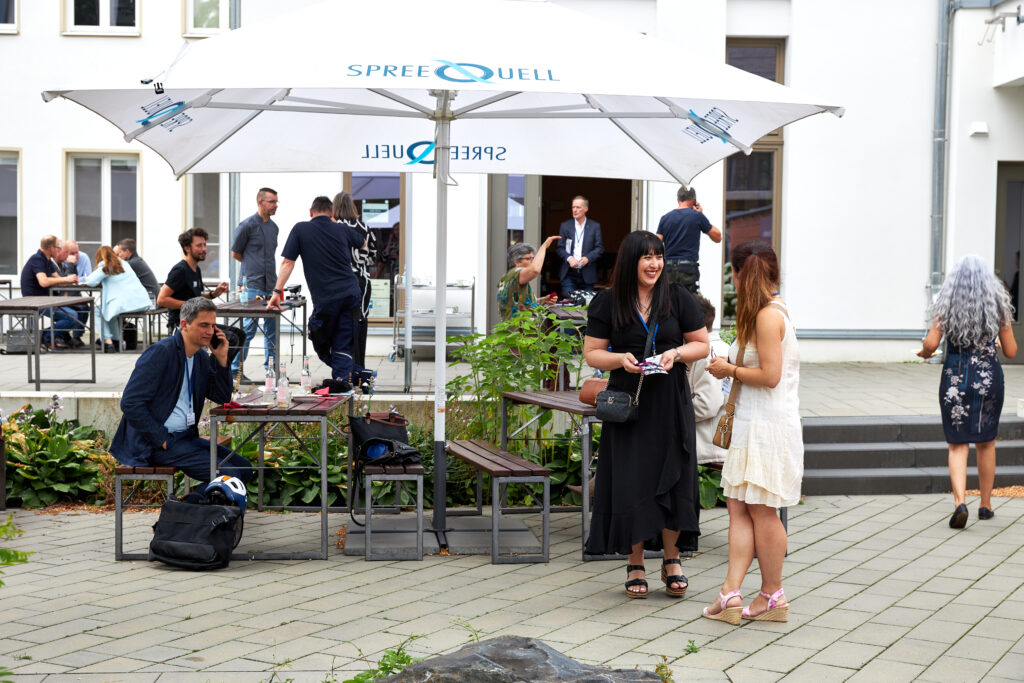Menschen im Außenbereich einer Veranstaltung im Gespräch. Zwei Frauen lachen, andere sitzen an Tischen oder bewegen sich zwischen Haus und Terrasse.