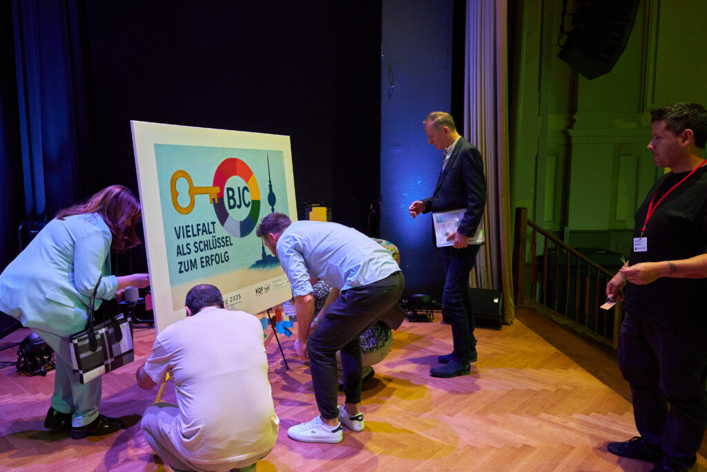 Mehrere Personen bringen bunte Zettel am partizipativen Kunstwerk auf der Bühne an. Das Plakat trägt die Aufschrift „Vielfalt als Schlüssel zum Erfolg“.