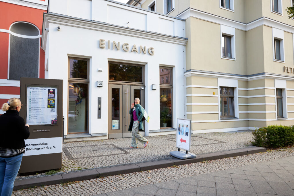 Eine Frau winkt beim Betreten des Peter Edel Bildungs- und Kulturzentrums in Berlin Weißensee, während eine andere Person das Veranstaltungsprogramm liest.