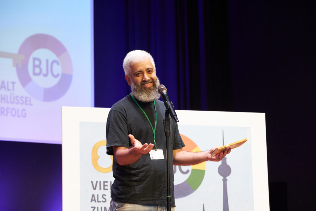 Aidin Halimi steht auf der Bühne und trägt beim Poetry Slam vor. Er spricht ins Mikrofon, gestikuliert und hält ein Notizbuch in der Hand.