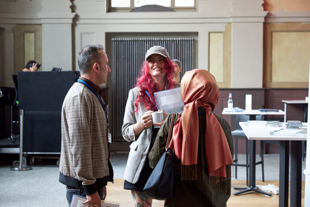 Drei Personen im Gespräch bei einer Veranstaltung. Eine Frau mit rotem Haar lacht, eine Frau mit Kopftuch und ein Mann hören zu.